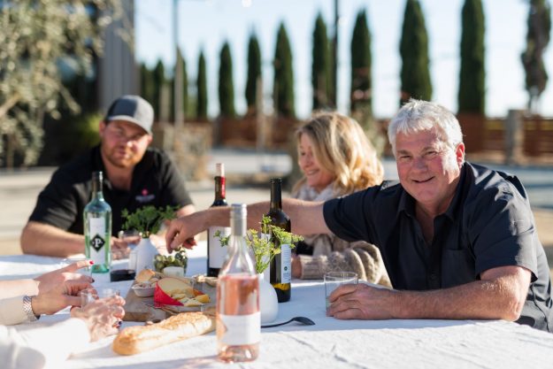 proprietor, Rod Schatz, smiling and sitting at a table with man and woman with wine, bread and cheese on a white tablecloth