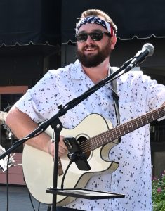 photo of mitchel dill in a white shirt with American Flag bandana on his head and holding a white guitar with a microphone stand in front
