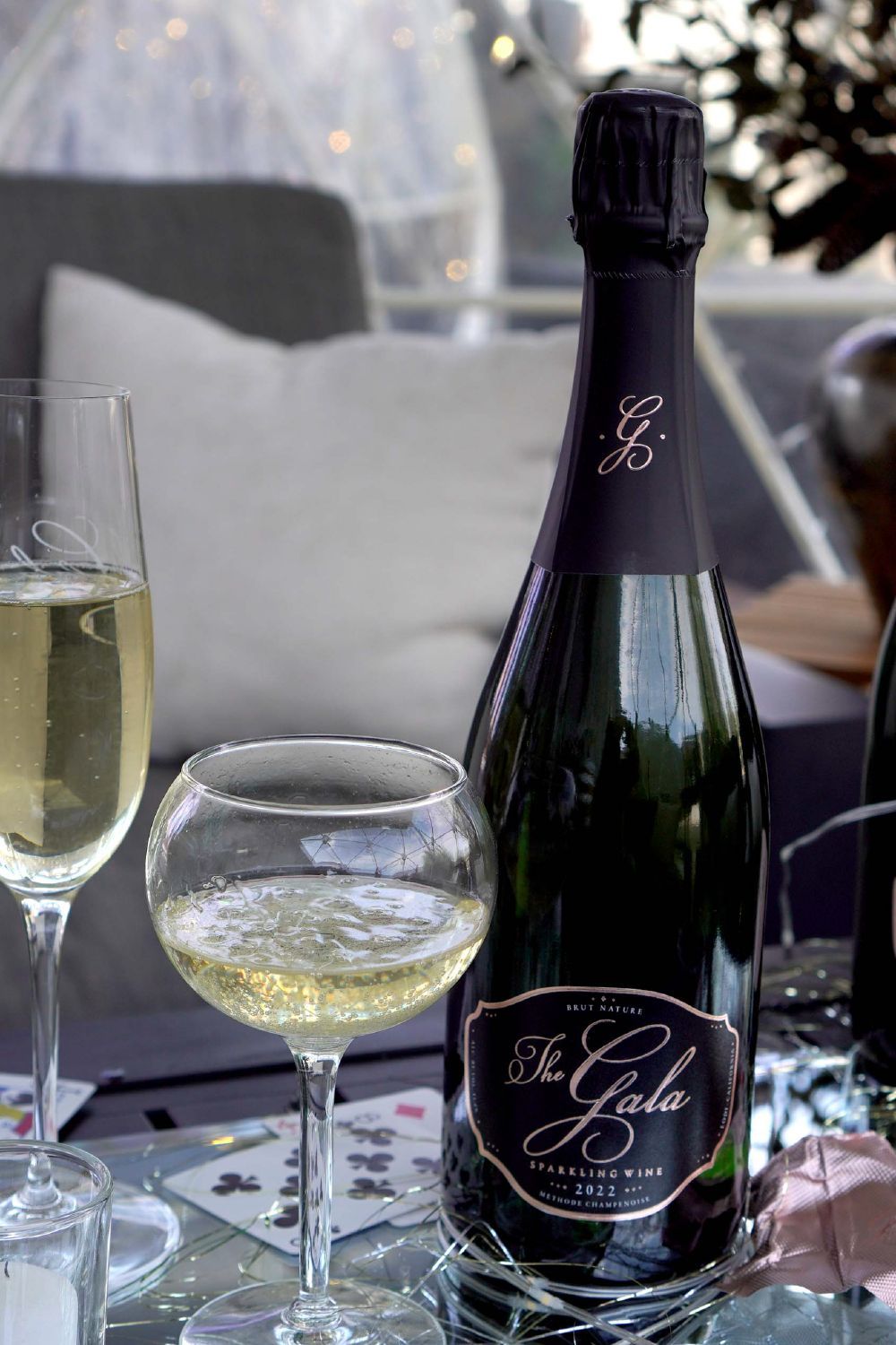 The Gala Sparkling Suite: a bottle of The Gala Brut Nature next to 2 glasses with wine - on a table with playing cards The Gala Sparkling Suite: a bottle of The Gala Brut Nature next to 2 glasses with wine - on a table with playing cards