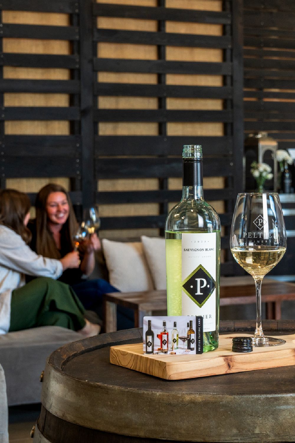 friends sitting on couch in the background, bottle of sauvignon blanc on barrel next to glass of wine and tasting card friends sitting on couch in the background, bottle of sauvignon blanc on barrel next to glass of wine and tasting card