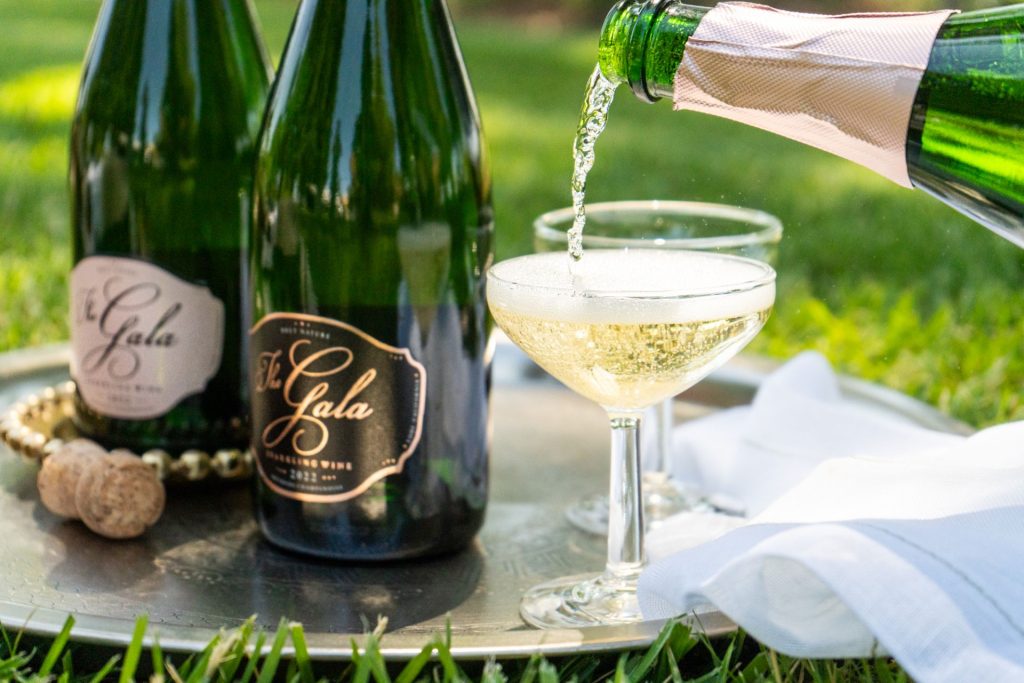 bottle of The Gala Sparkling wine being poured into a champagne glass next to other sparkling bottles bottle of The Gala Sparkling wine being poured into a champagne glass next to other sparkling bottles