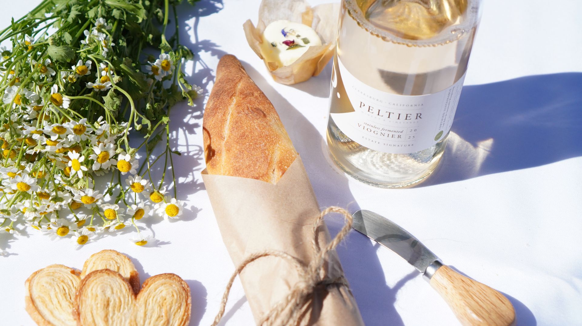 bottle of peltier viognier with small bunch of chamomile flowers, two palmier cookies, a baguette wrapped in parchment, a ball of butter and a cheese knife from above on a white tablecloth